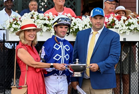 Coach Brad Cox receives the winner's trophy from Stephen Sanford's great-niece Suzi Salmen after the Mo Strike with jockey Florent Geroux on the winner's train after winning The Sanford's 107th run at Saratoga Racecourse Saturday, July 16, 2022 in Saratoga Springs, NY